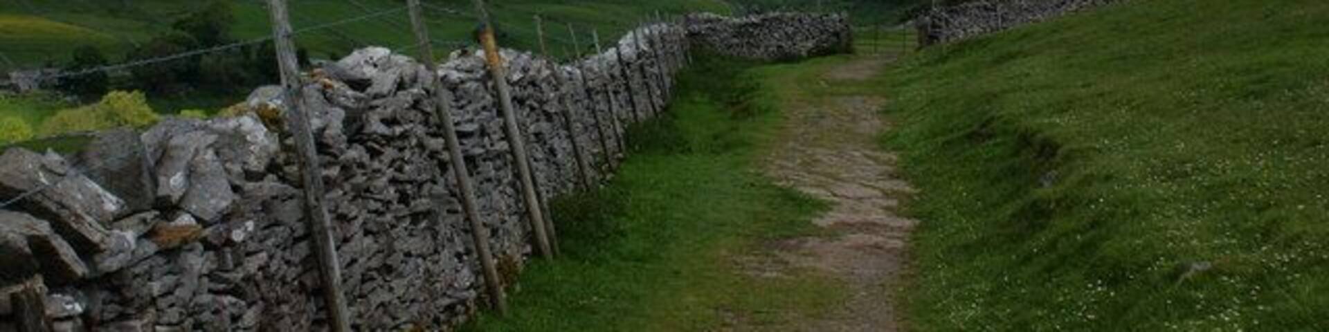 A stone wall, a grassy path, and rolling hills under a partly cloudy sky.