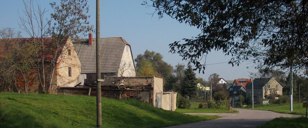 A rural road flanked by houses with red-tiled roofs and a grassy area.