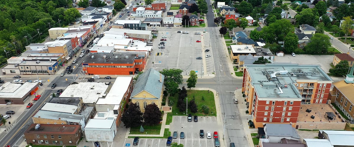 An aerial view of a town with a mix of commercial and residential buildings, a central parking lot, and surrounding greenery.