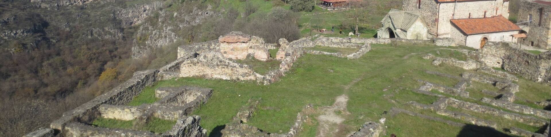 An ancient stone structure with ruins and a red-roofed building amidst a hilly landscape.