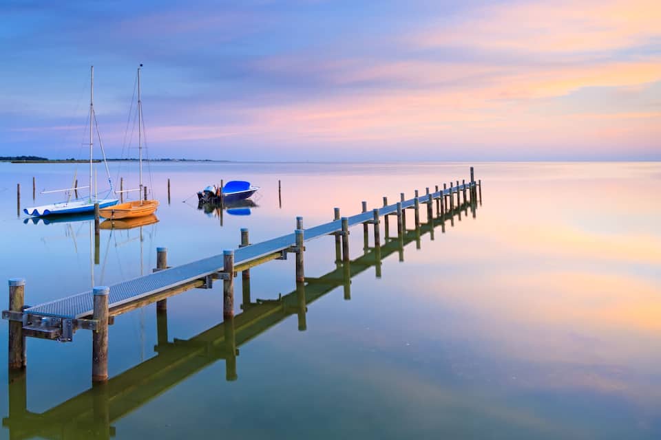 A calm lake with a wooden pier extending into the water, surrounded by boats and a clear sky.