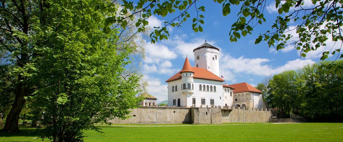 A castle with a red-tiled roof, surrounded by a stone wall and lush green trees.
