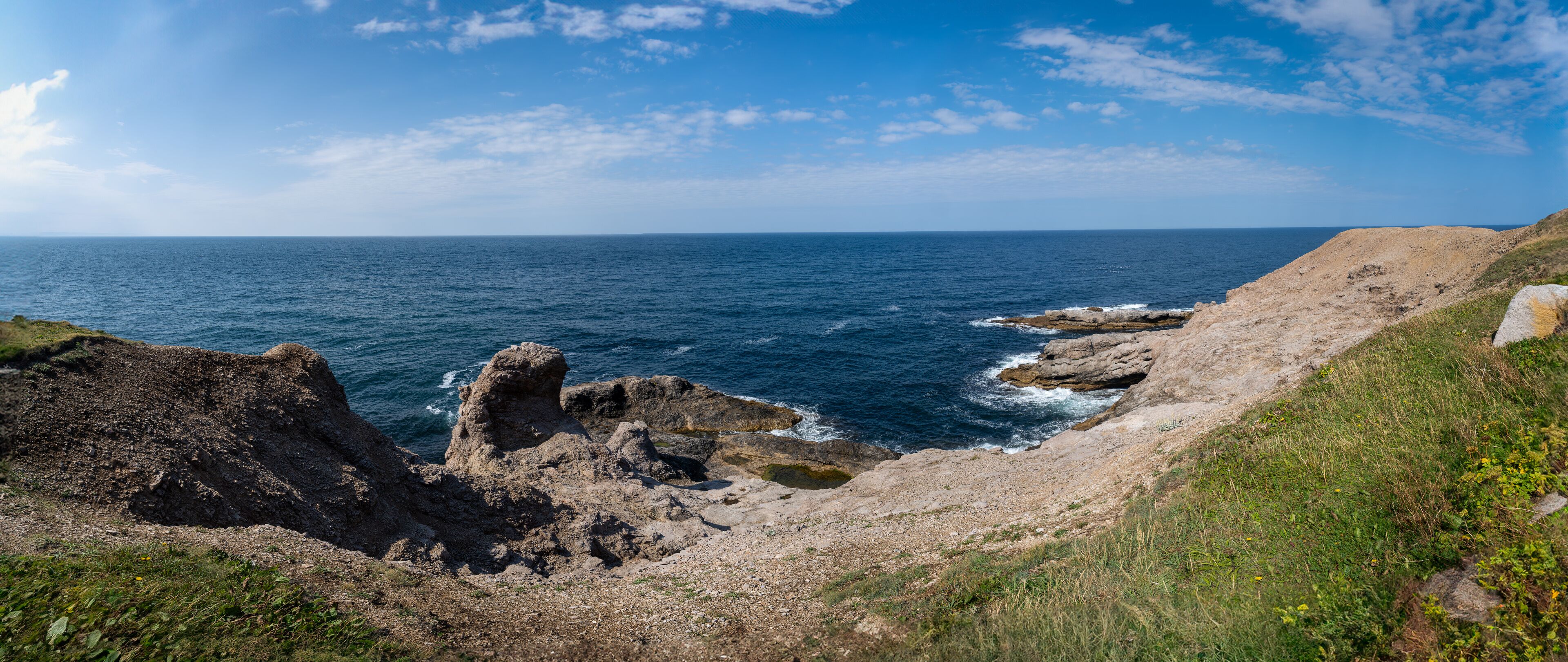 A coastal landscape with rocky cliffs, a clear blue sea, and a partly cloudy sky.