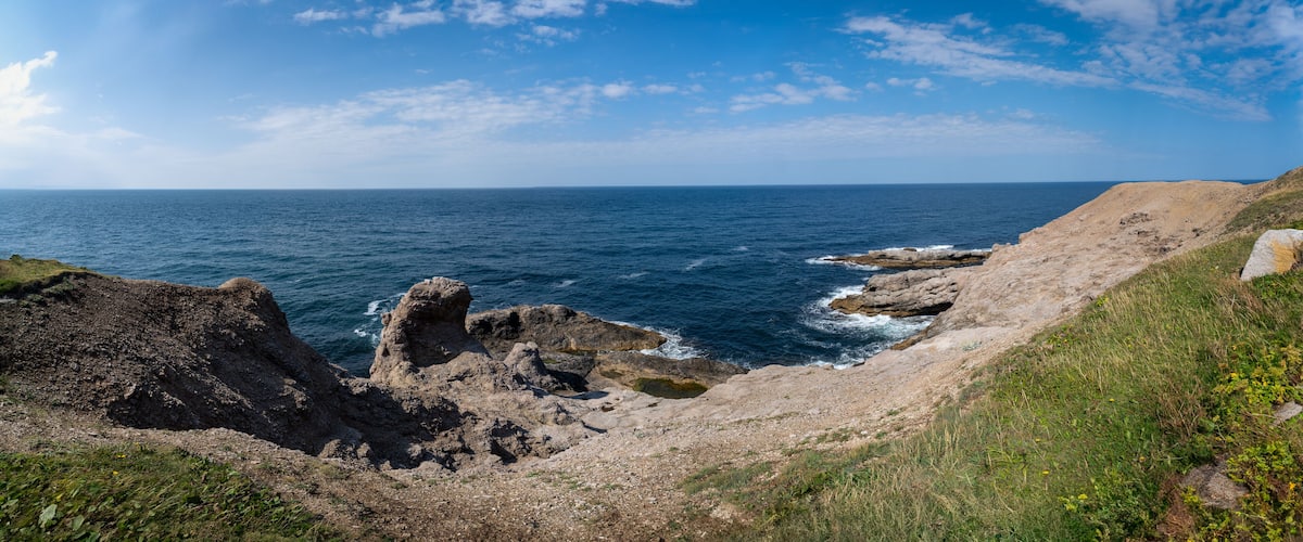 A coastal landscape with rocky cliffs, a clear blue sea, and a partly cloudy sky.