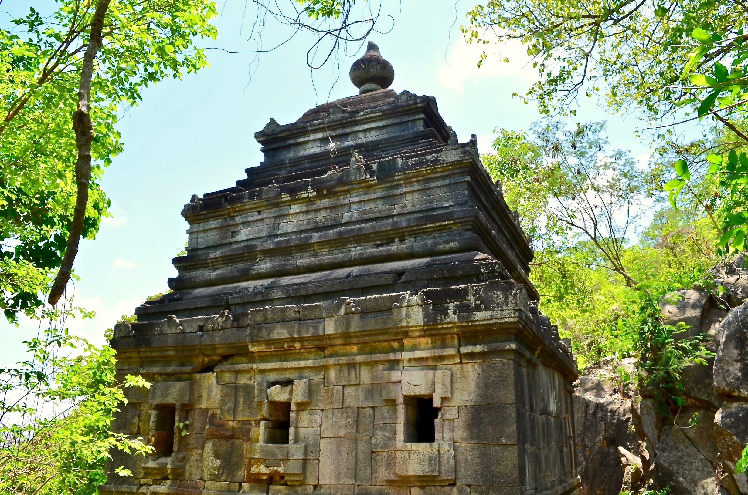 Um templo de pedra com telhado escalonado, rodeado por vegetação exuberante e árvores.