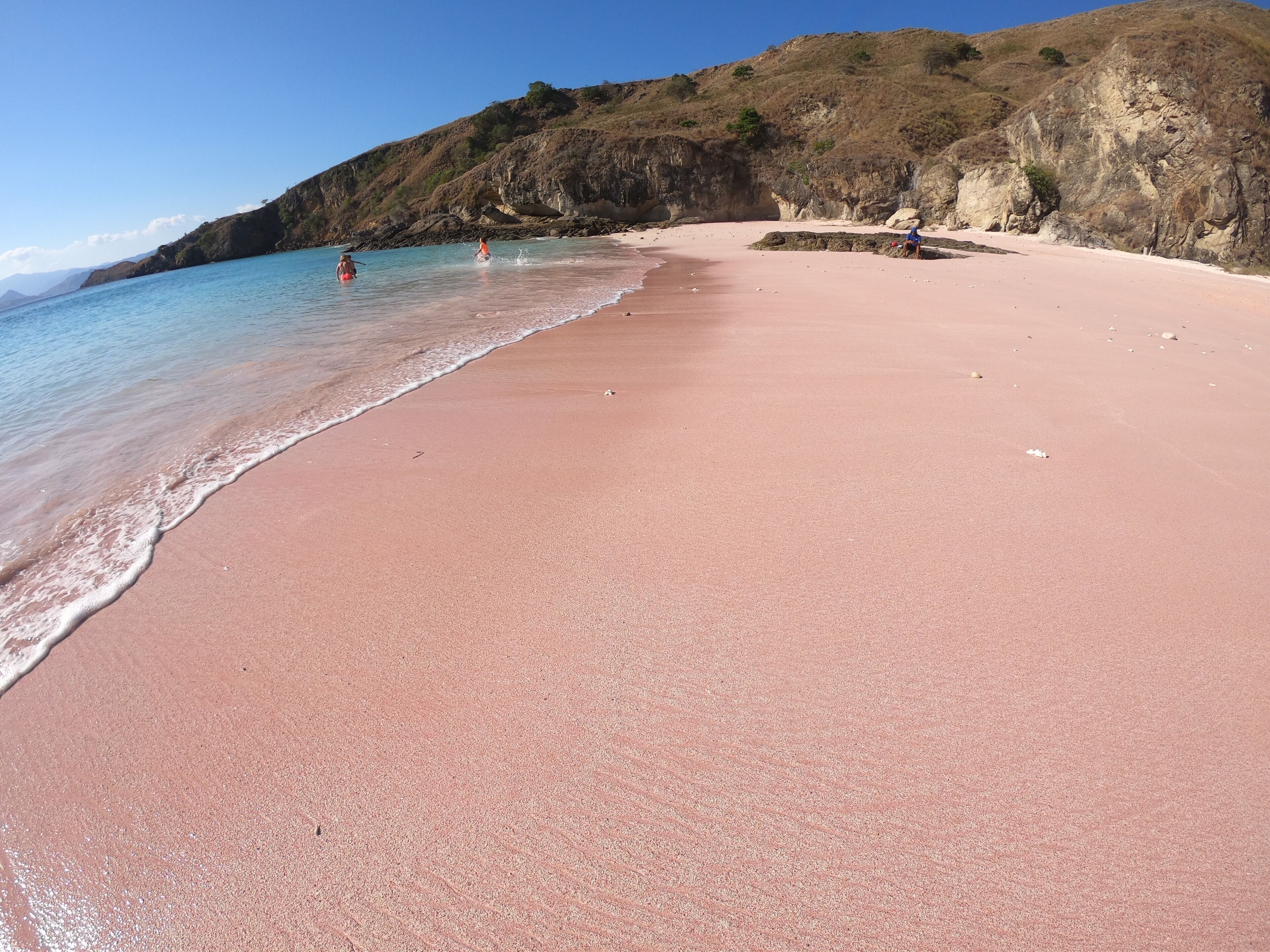Uma praia com areia rosa e águas azul-claras, rodeada por falésias rochosas.