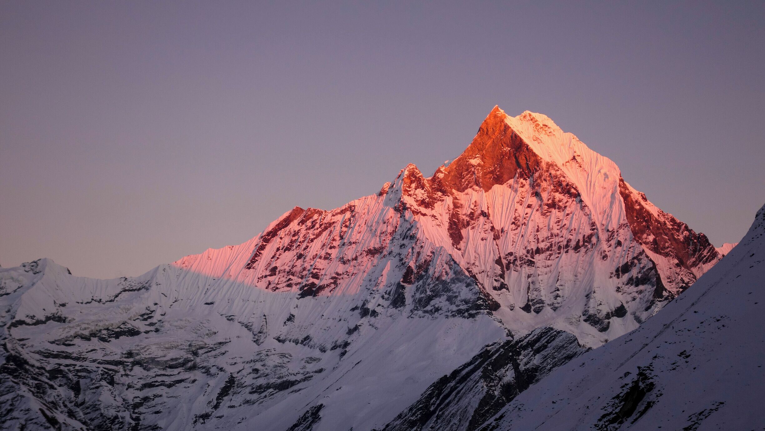 A snow-covered mountain peak with a reddish glow at its summit, surrounded by snow-covered slopes.