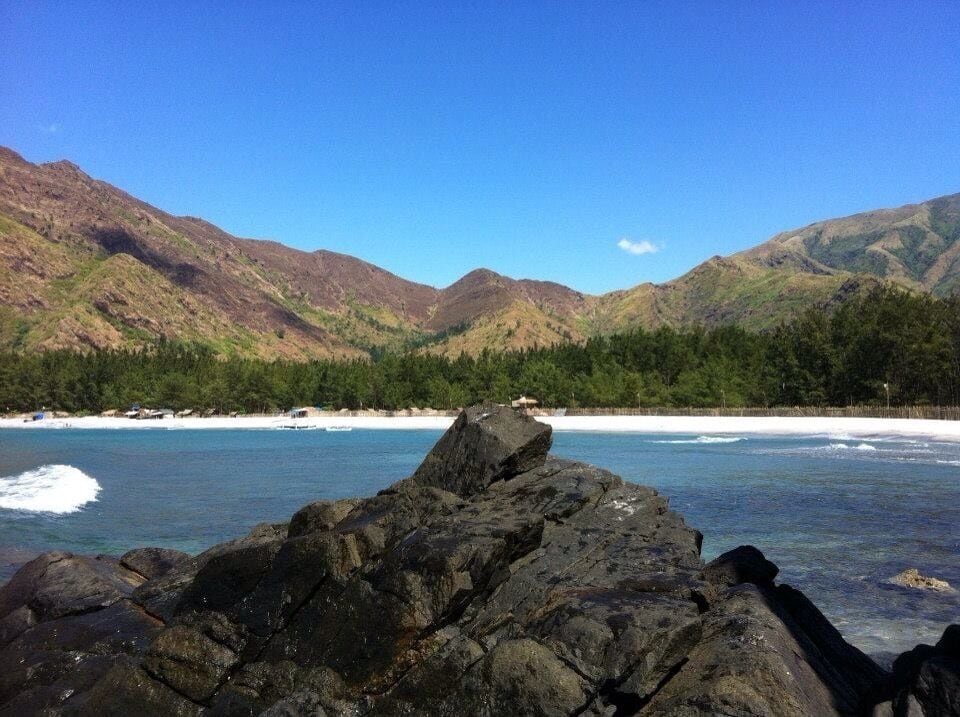 A rocky shoreline with clear blue water, a sandy beach, and forested mountains in the background.