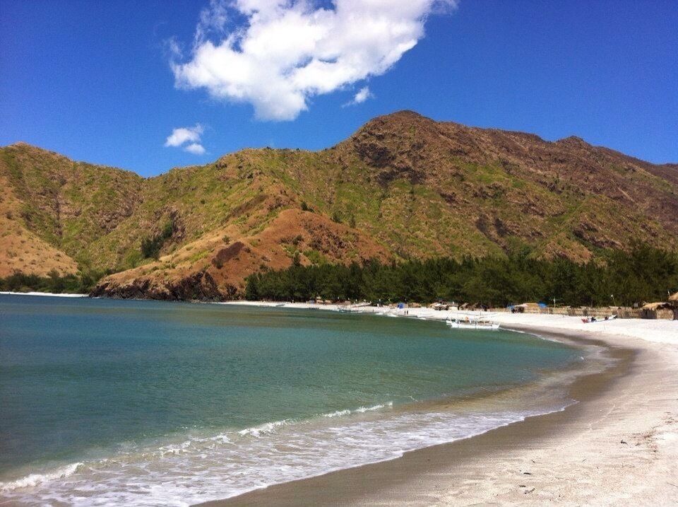 A beach with clear turquoise water, a sandy shore, and a mountain range in the background.