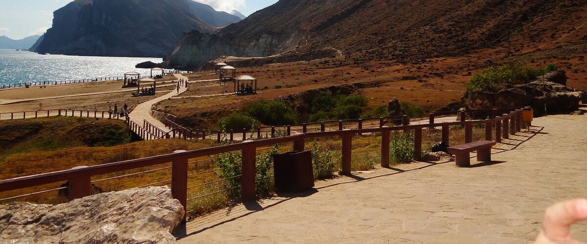 A coastal landscape with a rocky mountain, a beach, and a paved walkway.