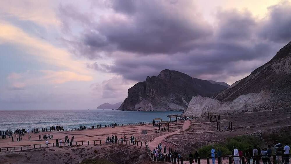 Eine Küstenlandschaft mit einem felsigen Felsen, einem Strand voller Menschen und einem klaren Himmel.