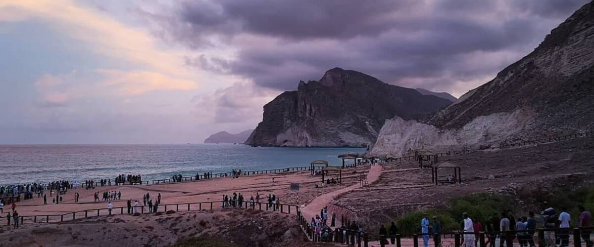 A coastal landscape with a rocky cliff, a beach with people, and a clear sky.
