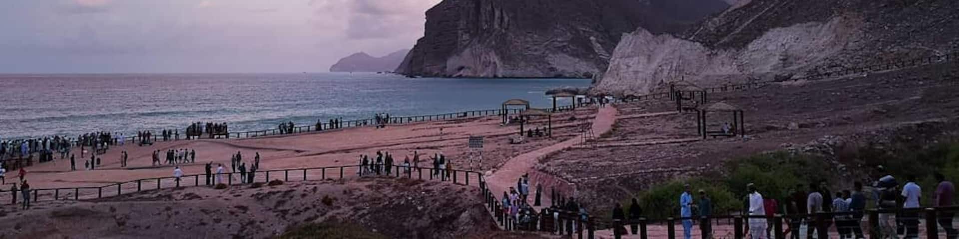 A coastal landscape with a rocky cliff, a beach with people, and a clear sky.
