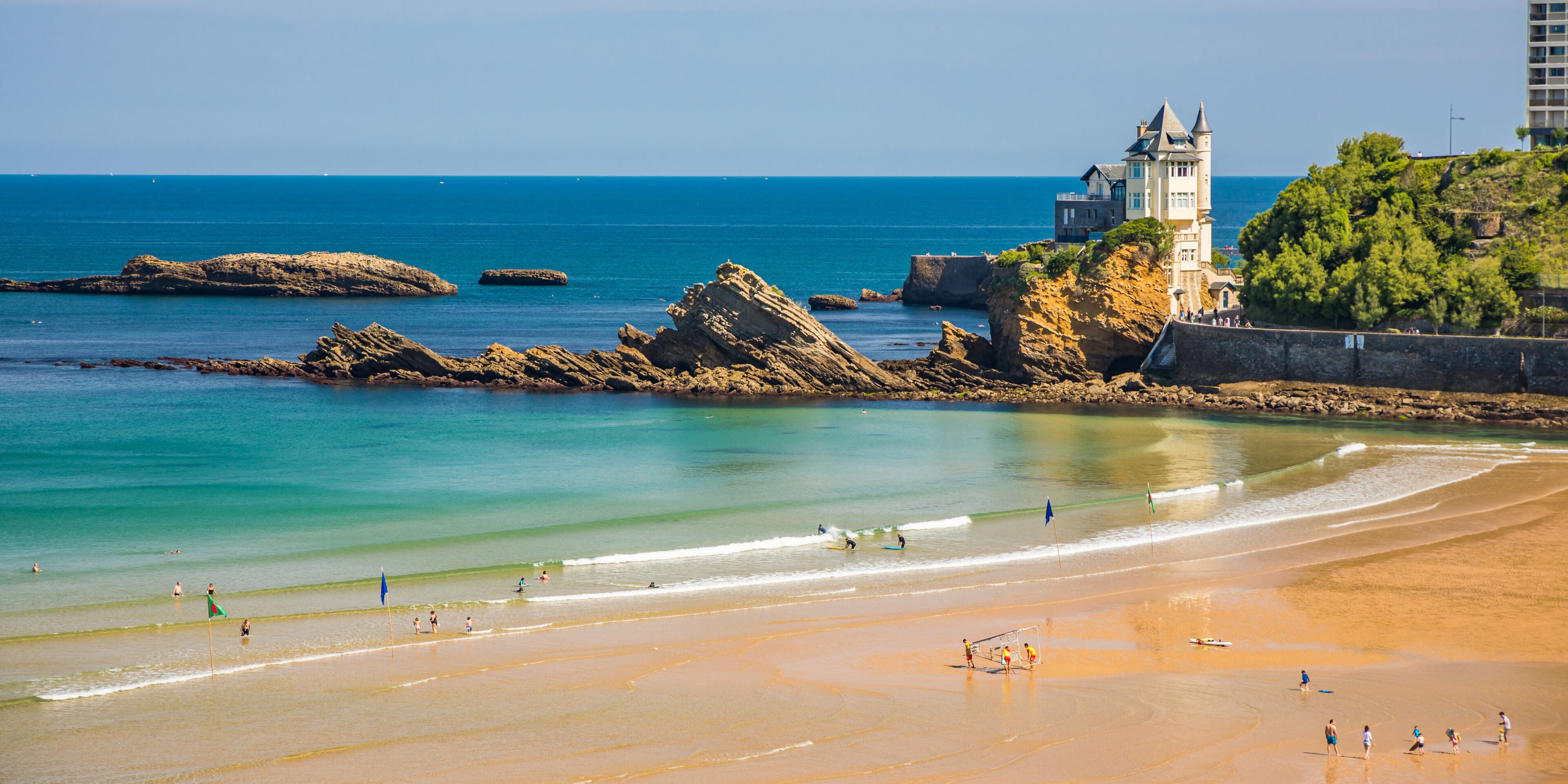 Uma praia costeira com águas claras, rochedos e uma estrutura semelhante a um castelo numa colina.