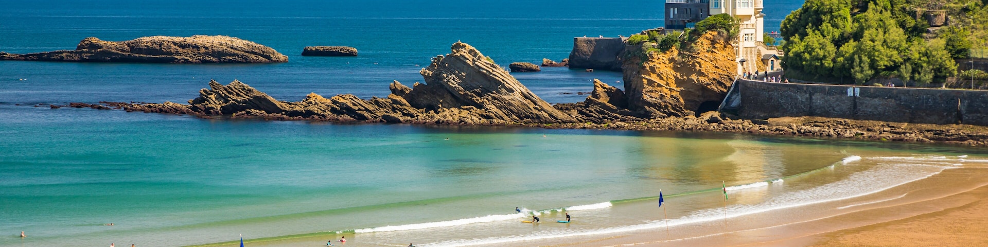 Uma praia costeira com águas claras, rochedos e uma estrutura semelhante a um castelo numa colina.