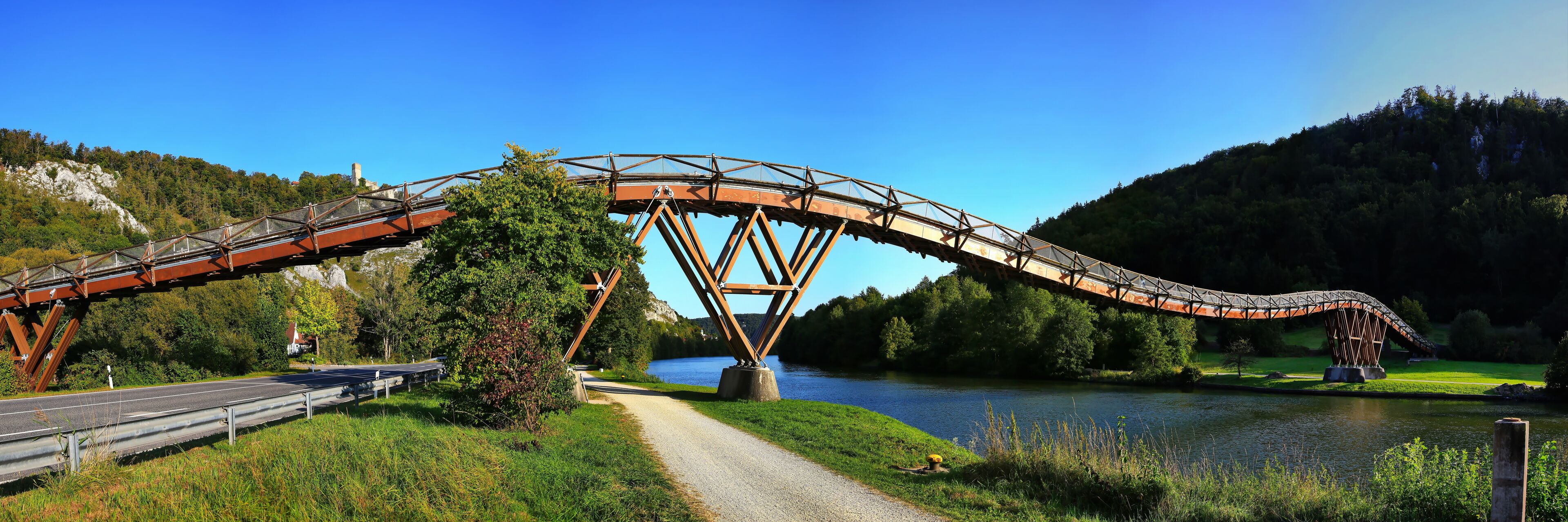 A steel bridge over a river, surrounded by greenery and hills.
