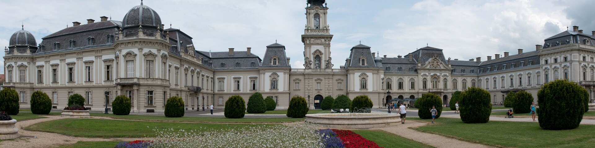 Ein prächtiges, historisches Gebäude mit einem zentralen Turm, umgeben von gepflegten Gärten und blauem Himmel.