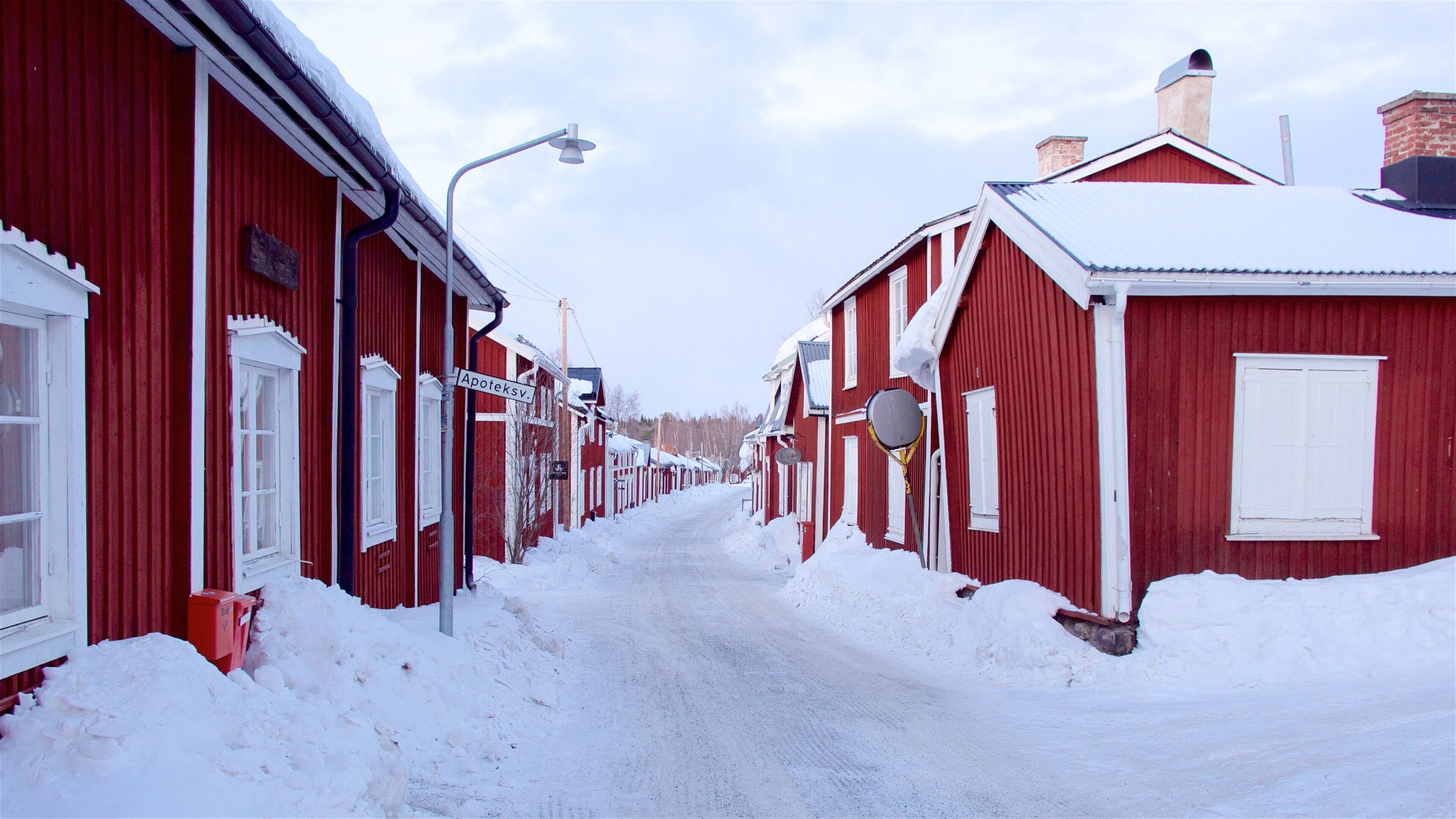 A snow-covered street lined with red buildings, a street lamp, and a sign reading "Apoteksv."