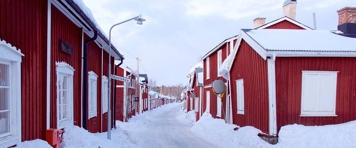 A snow-covered street lined with red buildings, a street lamp, and a sign reading "Apoteksv."