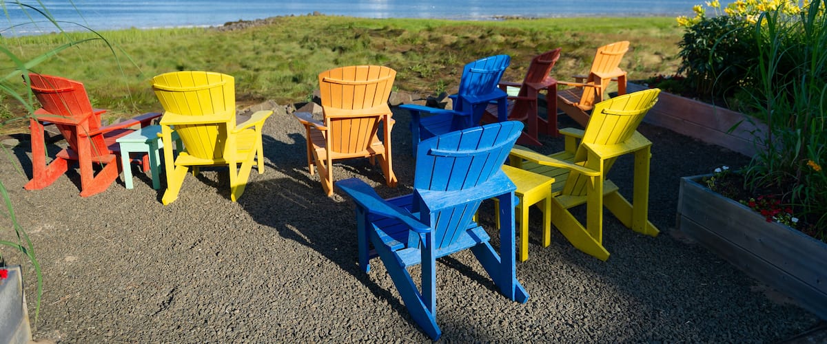 A gravel area with colorful Adirondack chairs and a view of a river with a boat in the distance.