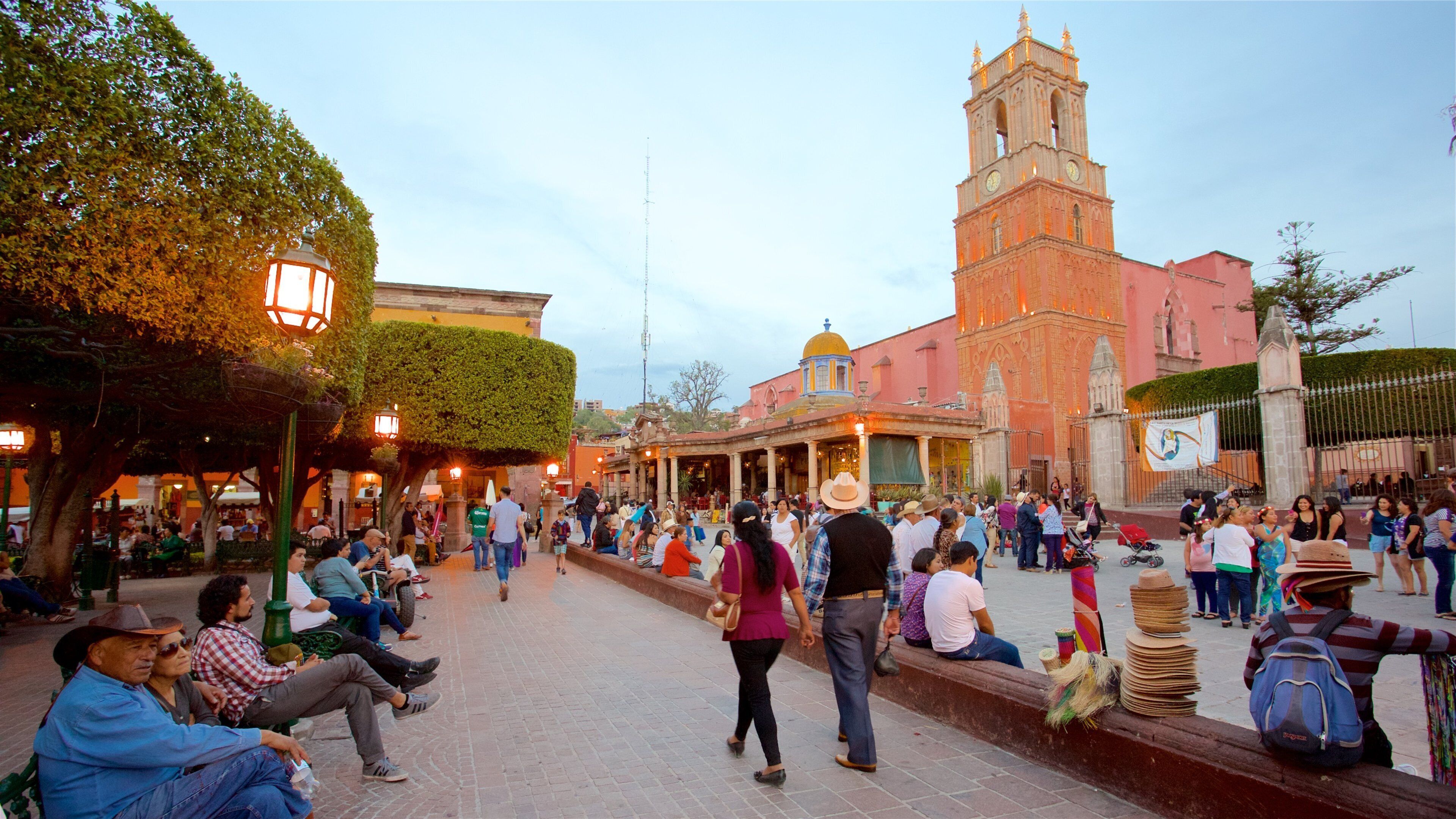 A bustling square with a prominent church tower, people walking, and a crowd gathered near a building with a yellow dome.