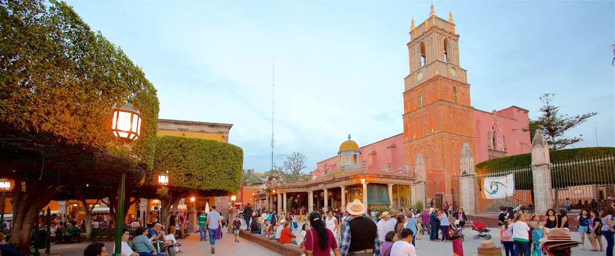 A bustling square with a prominent church tower, people walking, and a crowd gathered near a building with a yellow dome.