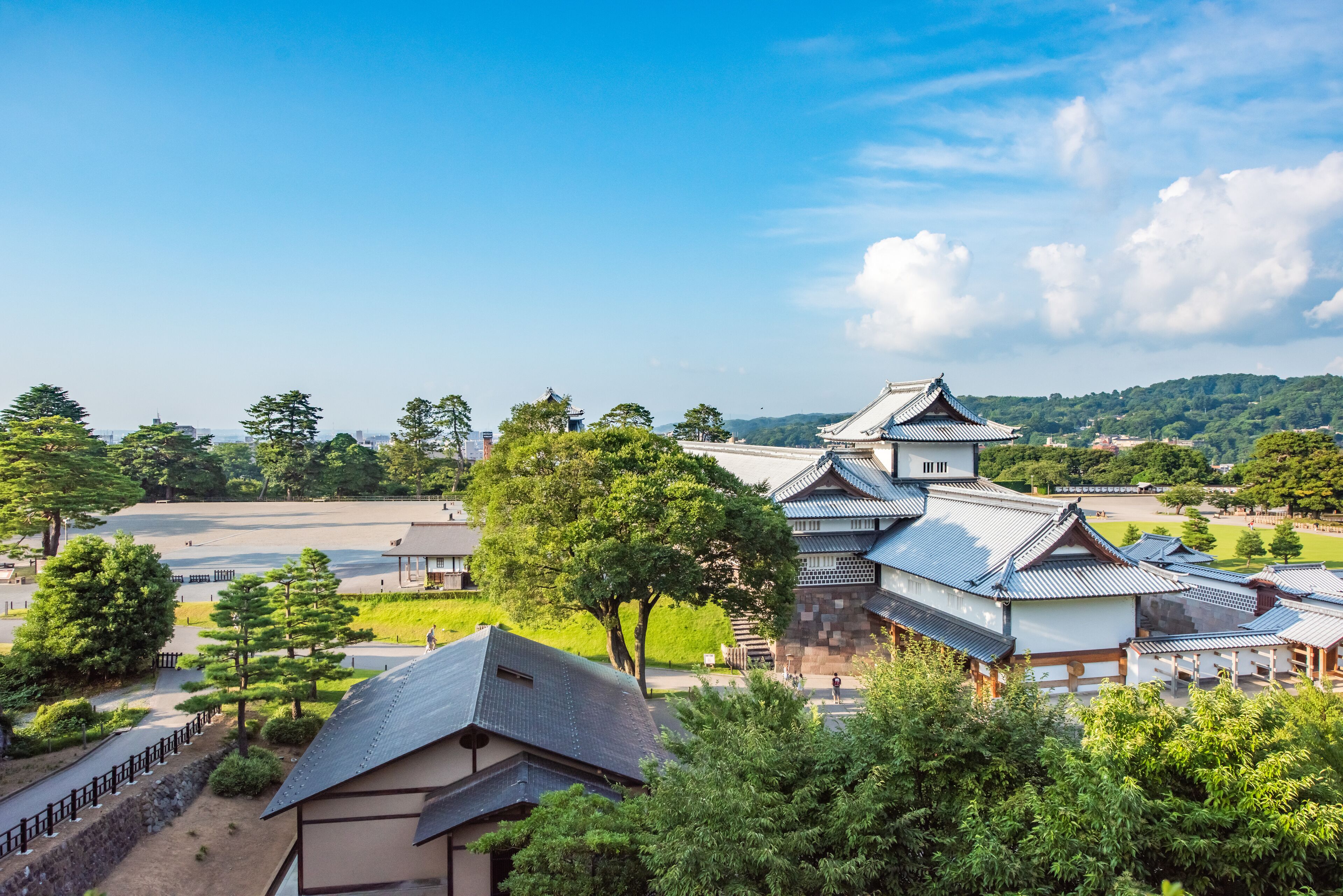 A traditional Japanese castle with multiple buildings, surrounded by lush greenery and a clear blue sky.
