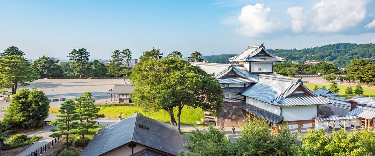 A traditional Japanese castle with multiple buildings, surrounded by lush greenery and a clear blue sky.