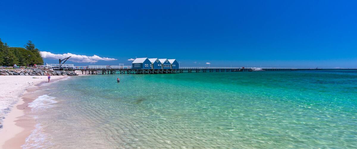 A beach with clear turquoise water, a wooden pier, and a row of blue buildings.