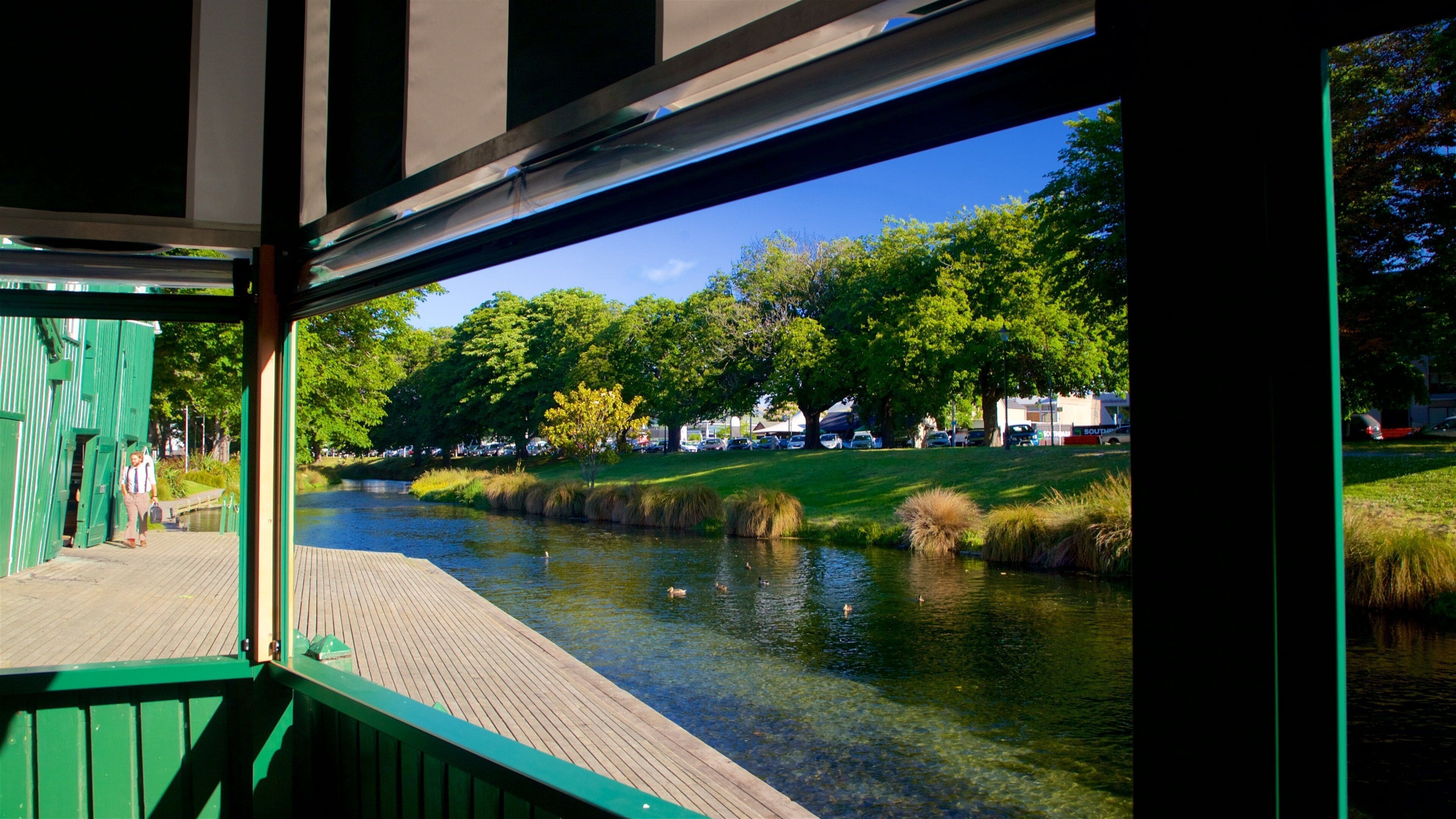 Um deck de madeira com vista para um rio calmo, onde patos nadam, cercado por árvores verdes e um céu azul límpido.