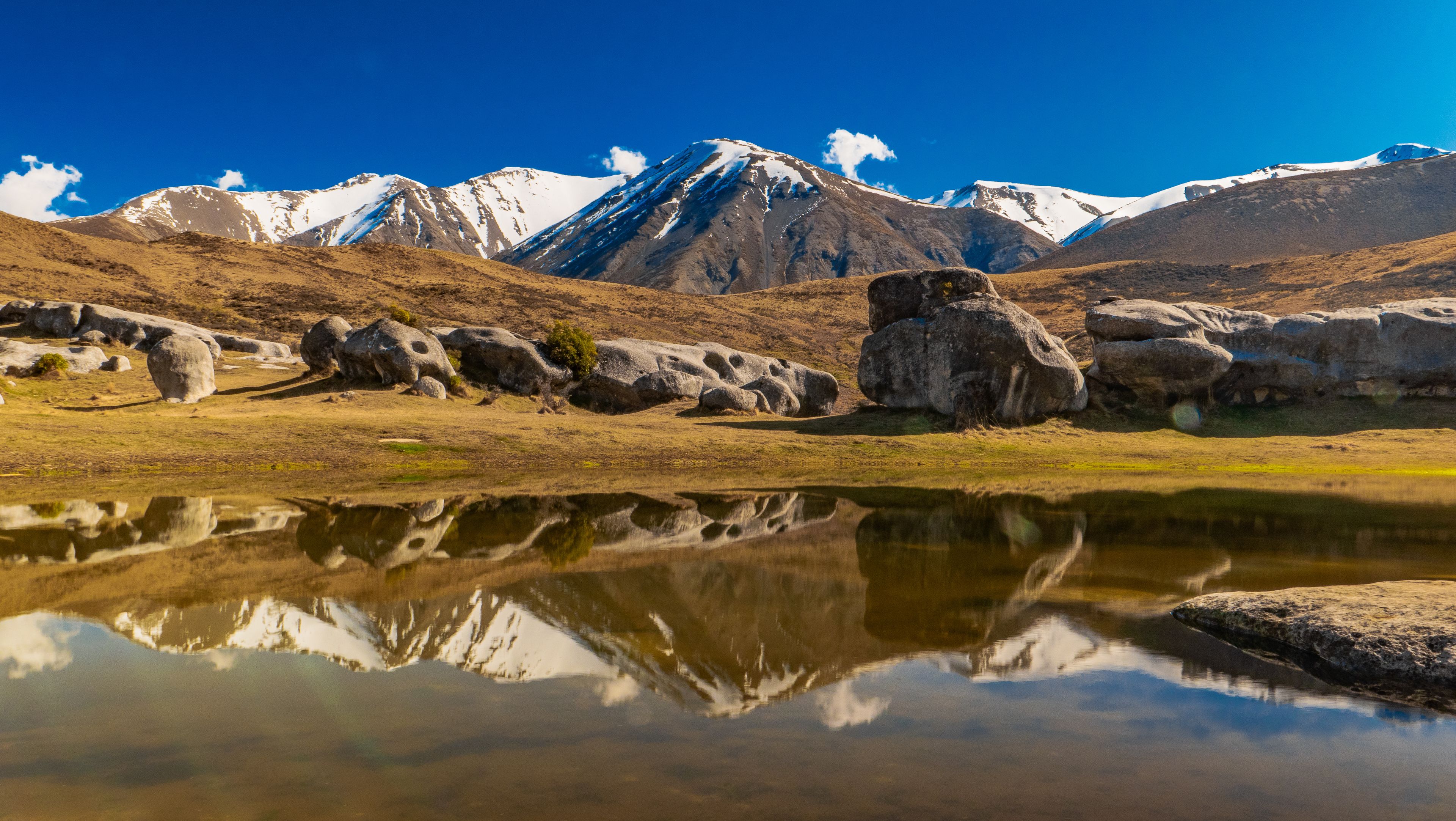 A mountain range with snow-capped peaks, a clear blue sky, and a calm body of water reflecting the landscape.