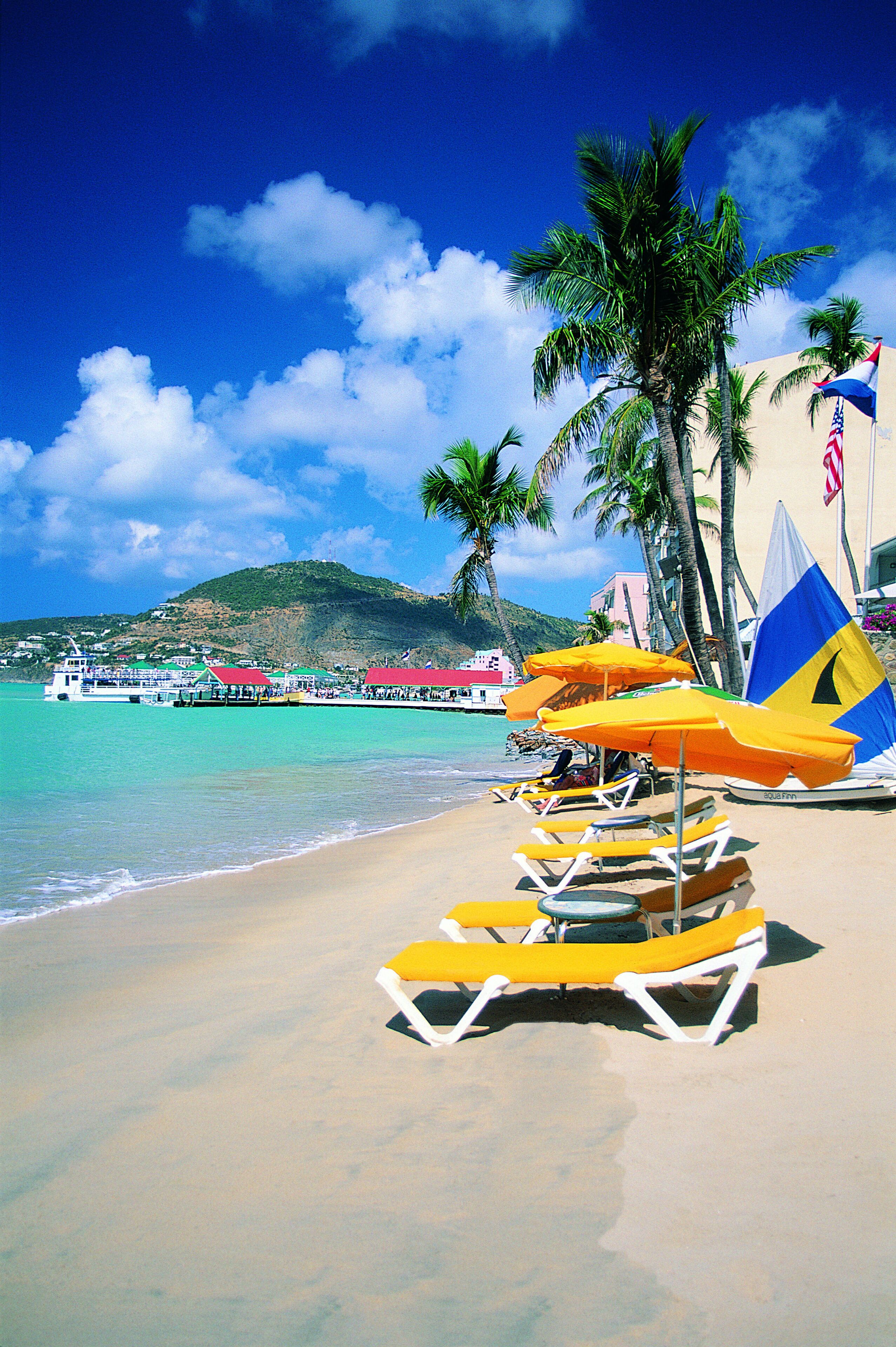 A beach with yellow lounge chairs, palm trees, a boat, and a pier.