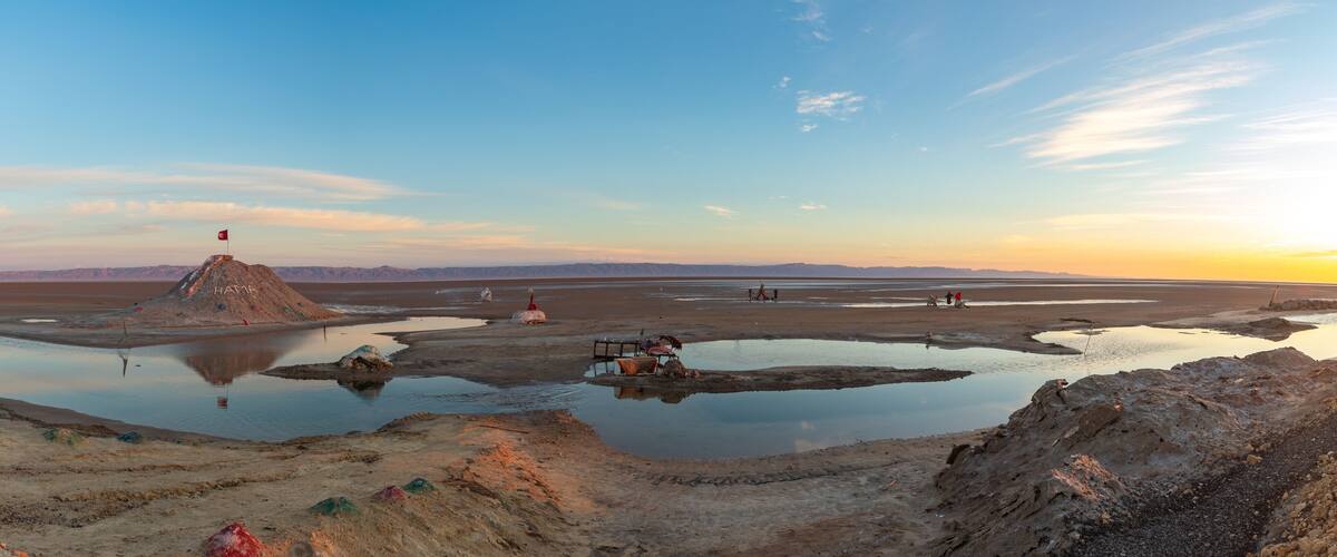 A salt flat with a flag on a hill, surrounded by water bodies and distant mountains.