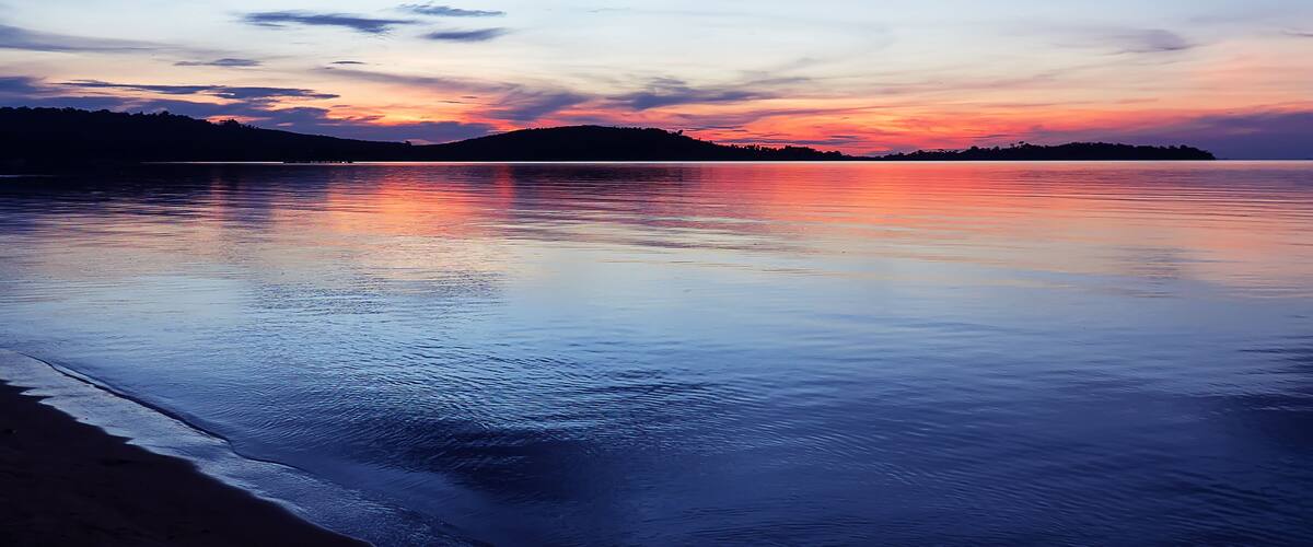 A calm beach at sunset with a silhouette of hills in the background.