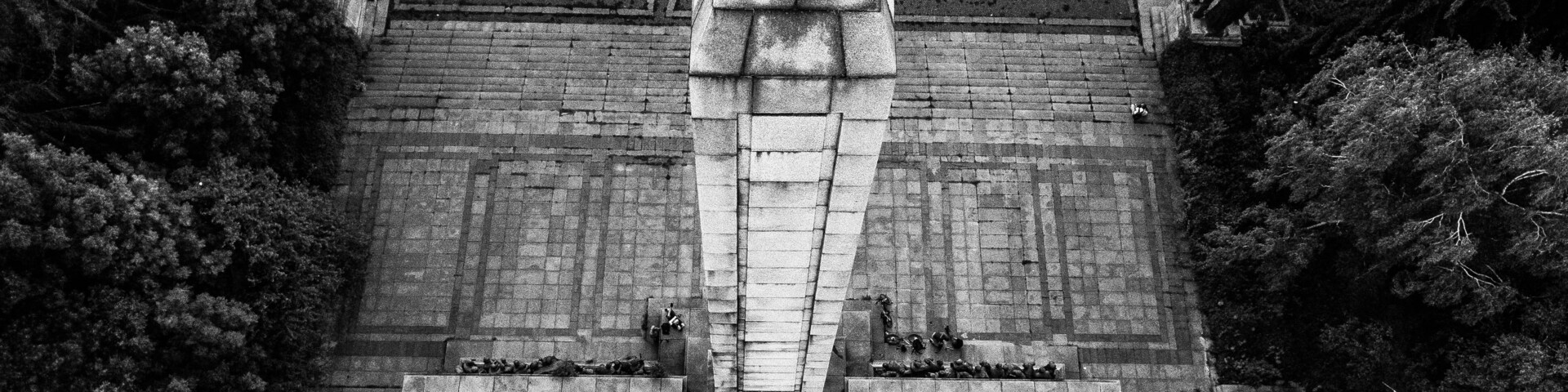 Um monumento de pedra com topo pontiagudo, ladeado por duas construções menores, com um céu claro e árvores ao fundo.