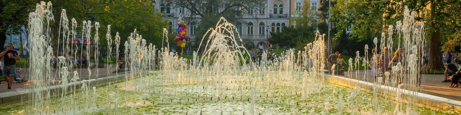 Uma fonte com jatos de água em um parque, com edifícios ao fundo.
