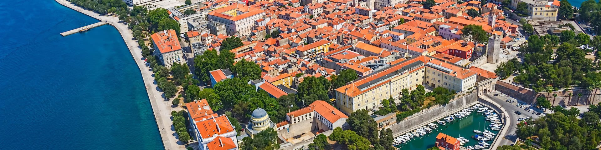 An aerial view of a coastal city with red-roofed buildings, a marina, and a prominent church.