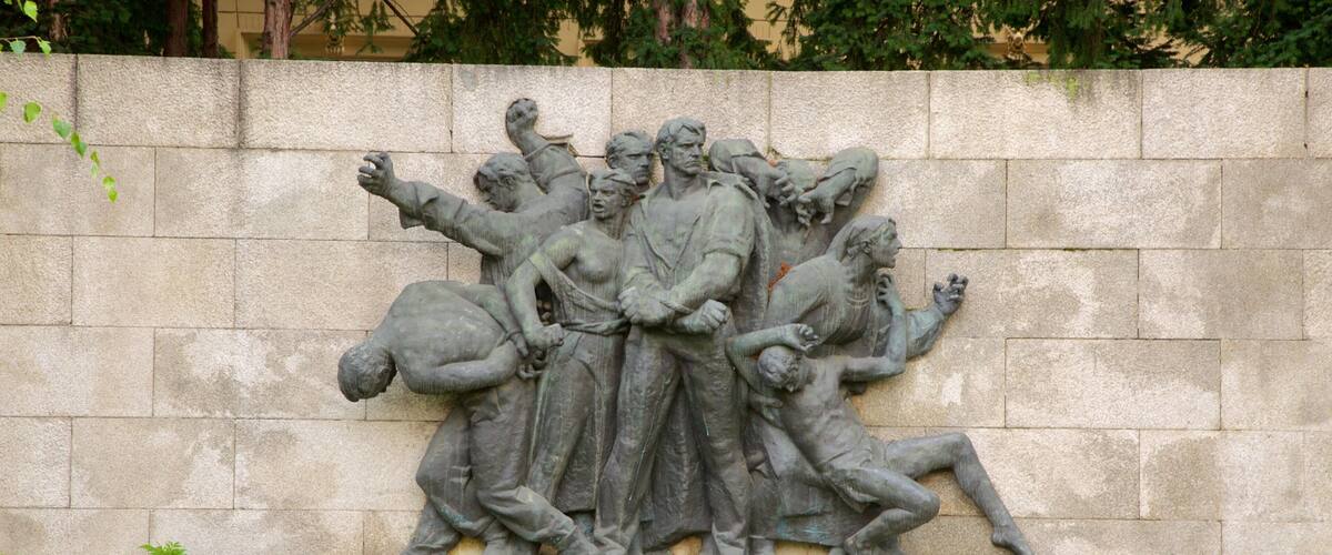 A stone relief sculpture of a historical scene with figures in various poses, set against a stone wall with greenery at the base.
