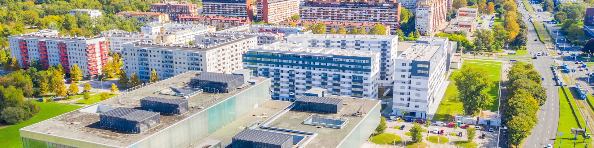 An urban landscape with modern buildings, a parking lot, and a green park area.