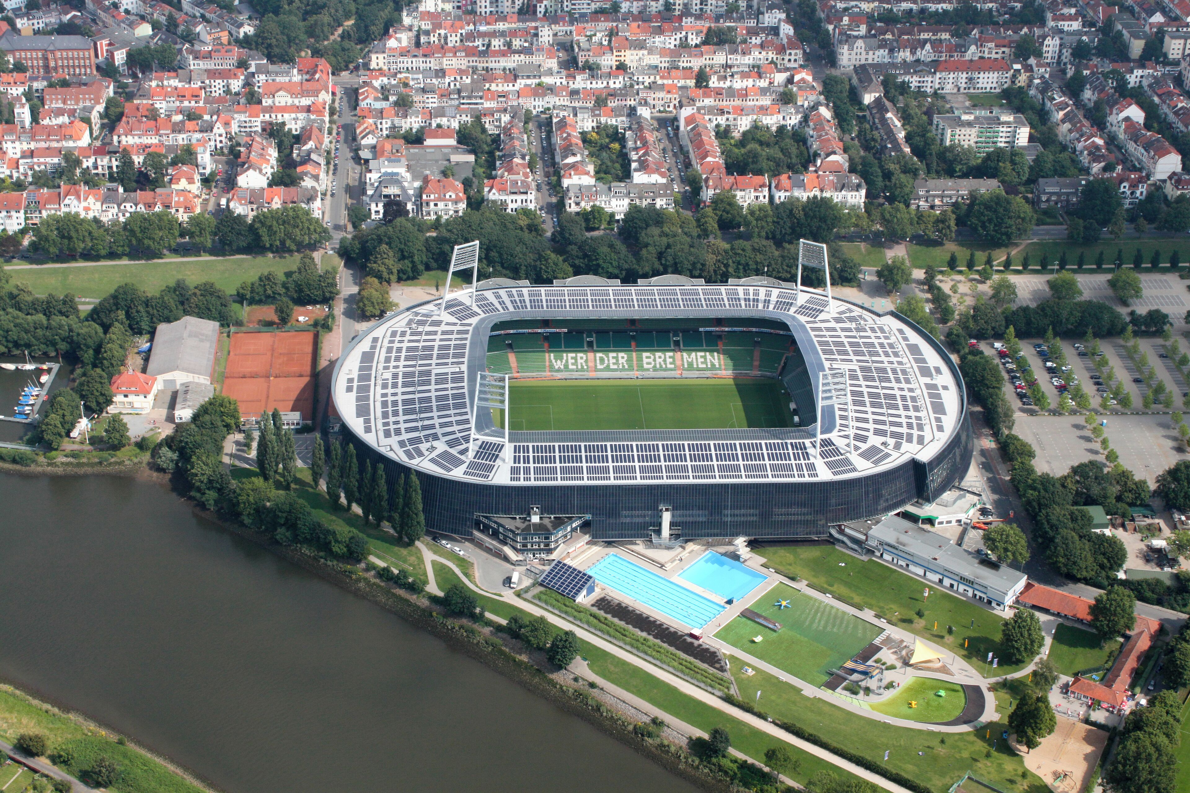 Vista aérea de un estadio con 'WER DER BREMEN' escrito en el campo, rodeado por un río, edificios residenciales y una piscina.