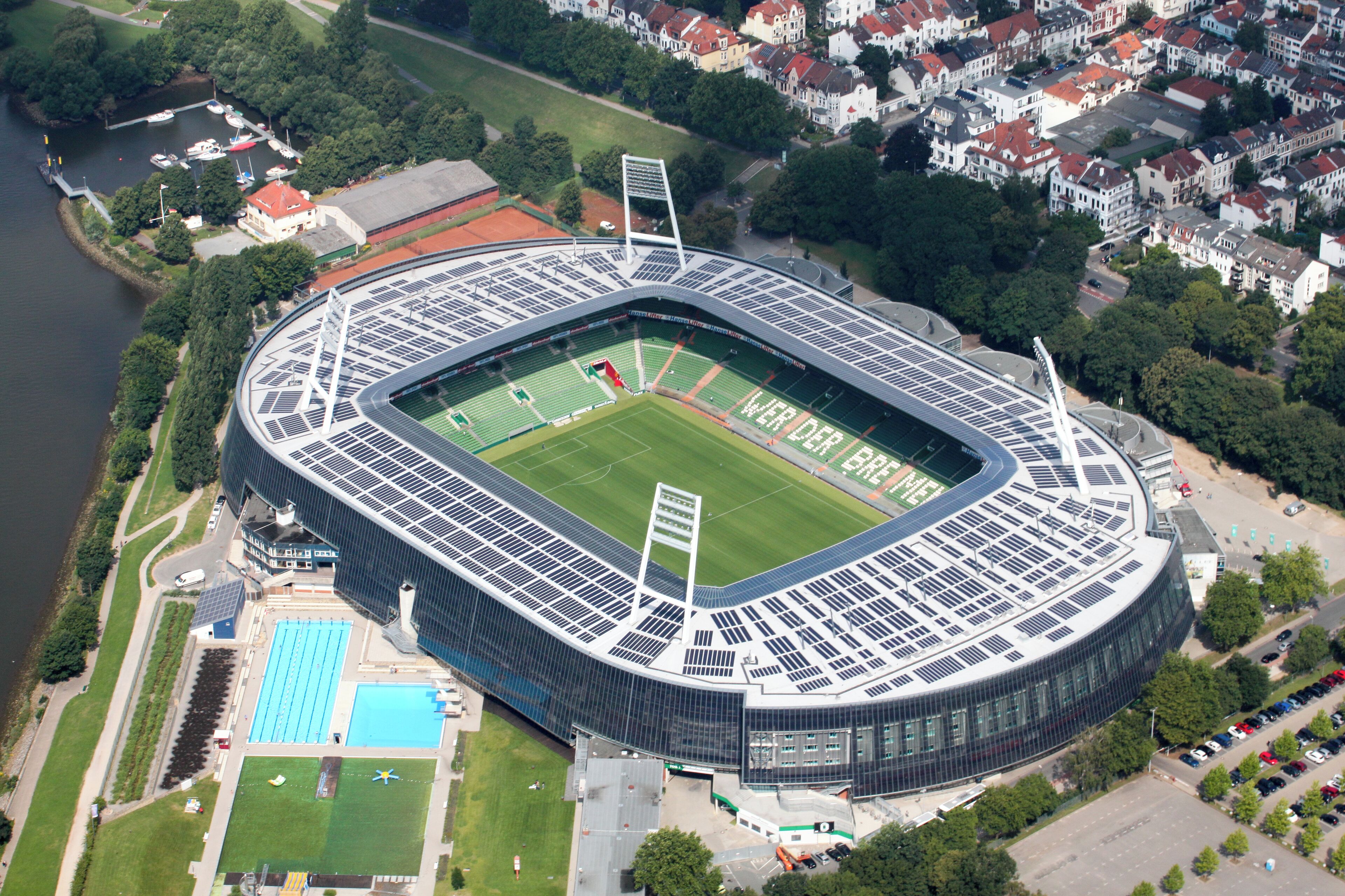 An aerial view of a modern stadium with a football field, solar panels, and a swimming pool.