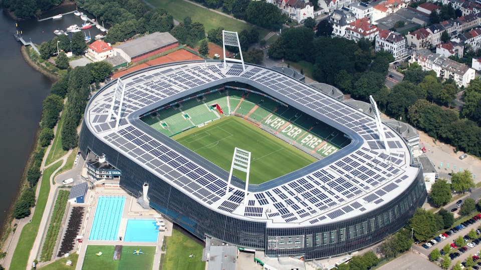 An aerial view of a modern stadium with a football field, solar panels, and a swimming pool.