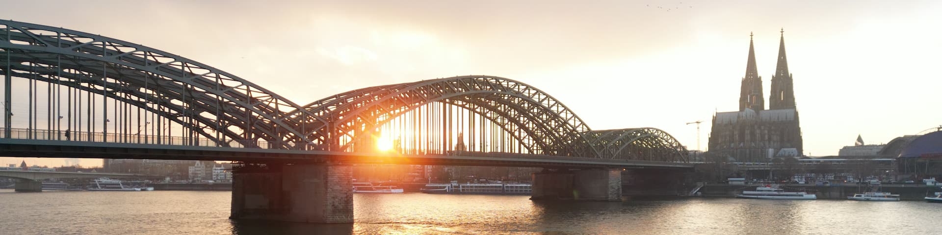 A steel bridge over a river with the sun setting behind it, and a cathedral with two spires in the background.