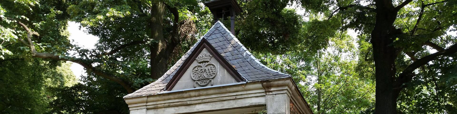 A small stone structure with a pointed roof and a coat of arms, surrounded by lush greenery and a white metal fence.