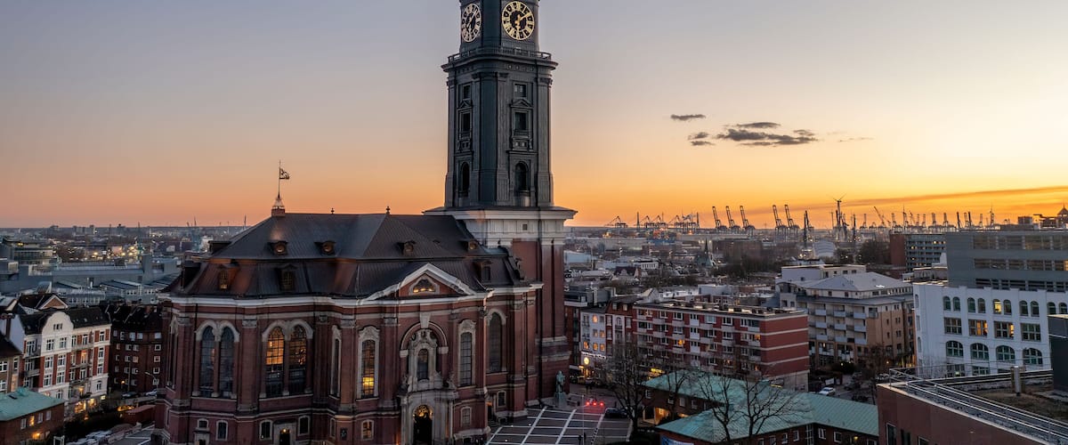 Ein historisches Gebäude mit einem Glockenturm, umgeben von moderner Architektur und einem Sonnenuntergangshimmel.