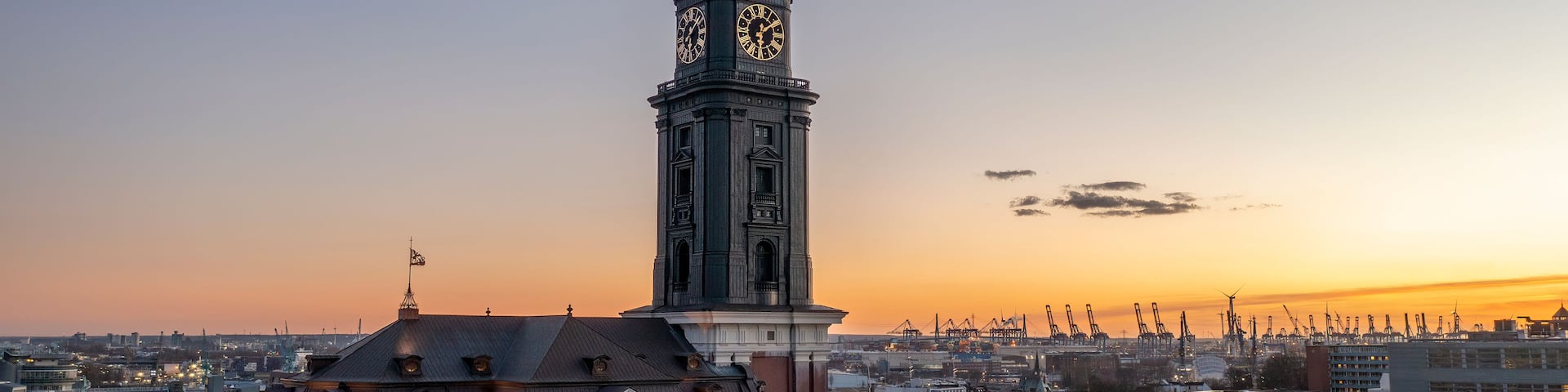 A historic building with a clock tower, surrounded by modern architecture and a sunset sky.