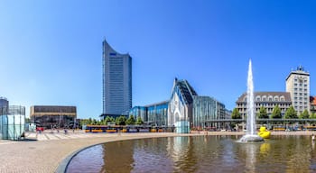 Una plaza urbana con una fuente, edificios modernos y una torre de reloj histórica.