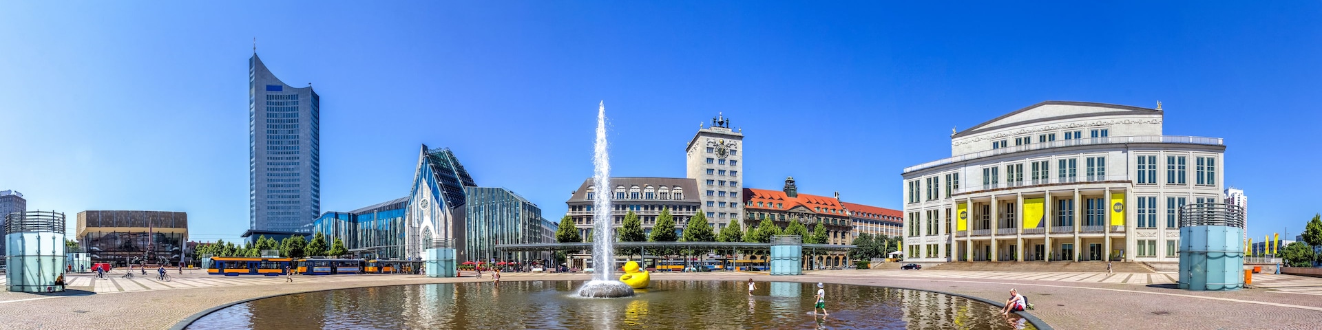 Uma praça urbana com uma fonte, edifícios modernos e um antigo campanário.