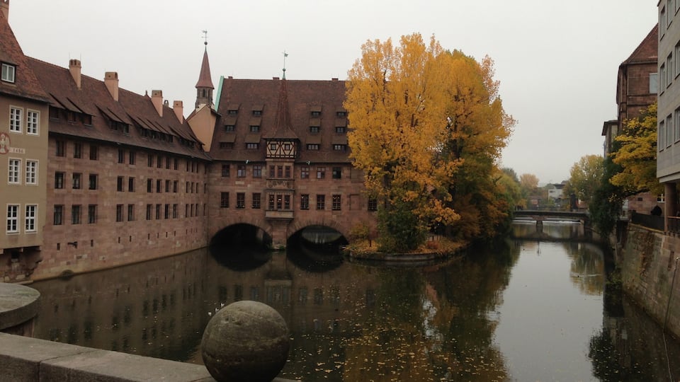 Um canal com uma esfera de pedra na margem, rodeado por edifícios históricos e uma árvore com folhas de outono.