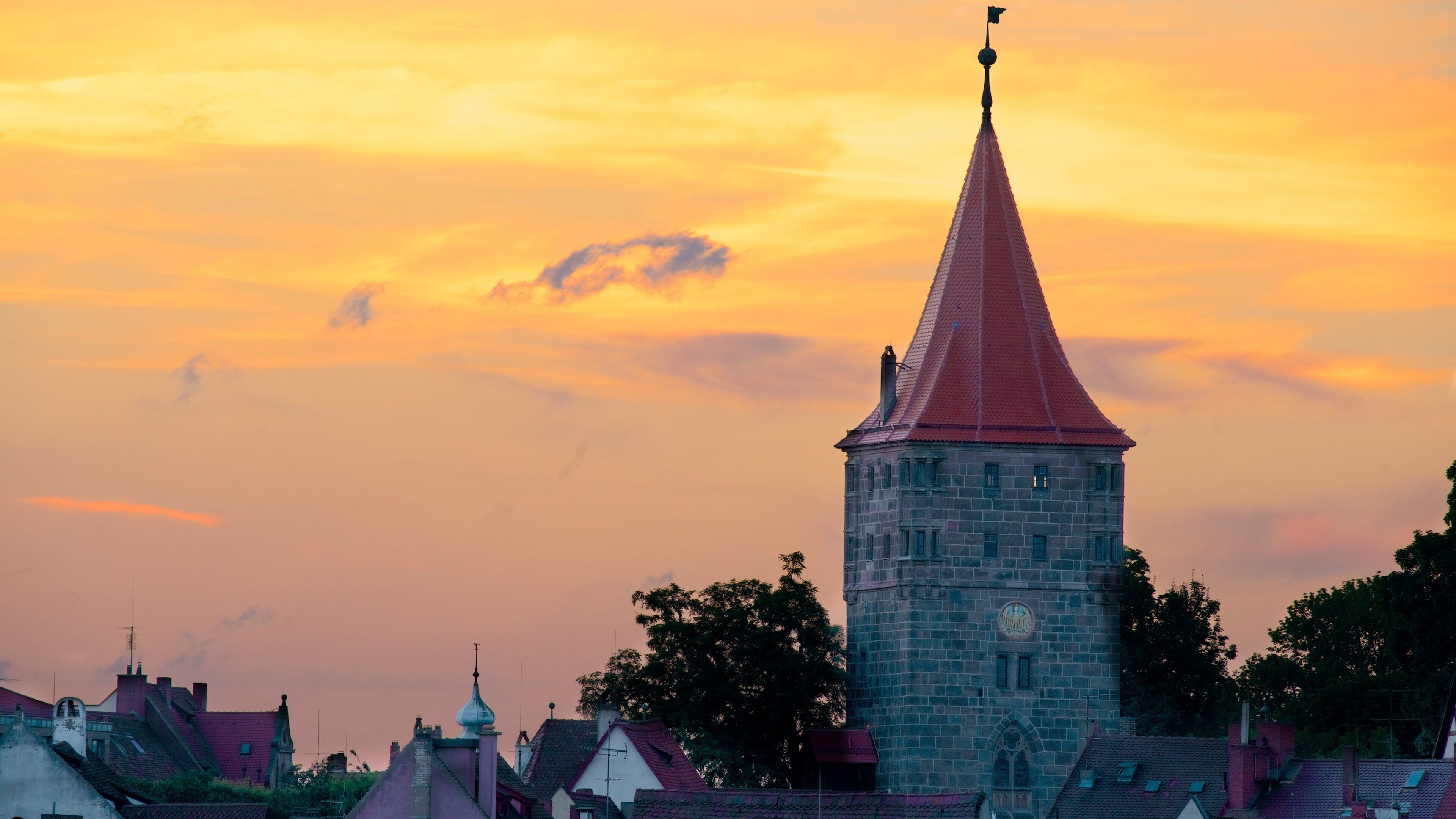 A tower with a red roof and a clock, surrounded by houses with red roofs, against a sunset sky.